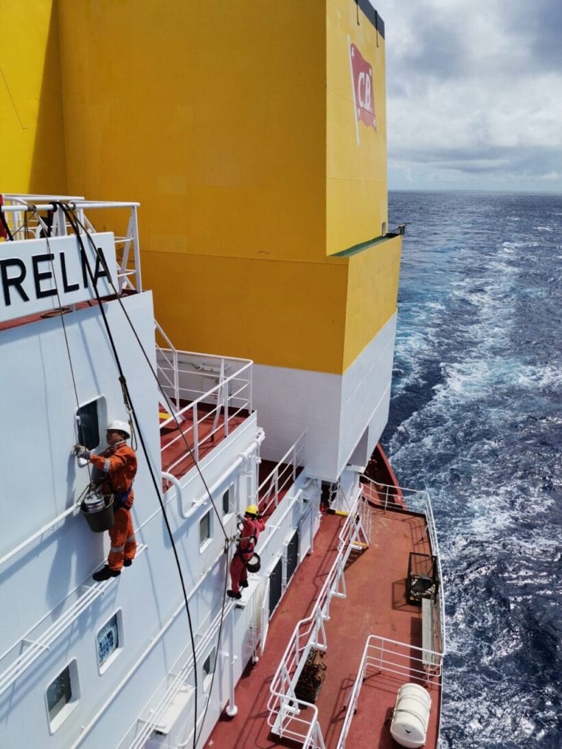 Deck crew painting on the side of the accommodation wearing safety harness and helmet.
