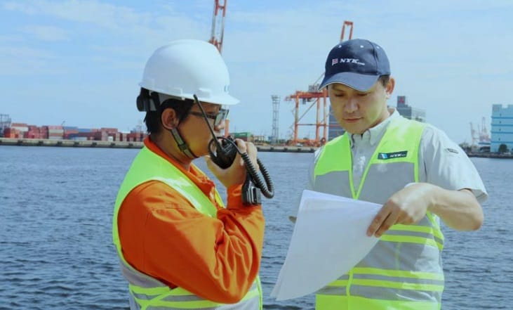 A crew wearing yellow coverall talking to someone using a walkie-talkie while his offer hands him a paper to read.