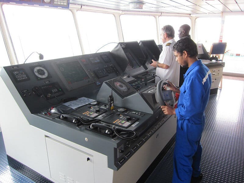 A helmsman wearing blue coverall steering a vessel while the Pilot and Ship Master give the commands during a maneuvering operation.