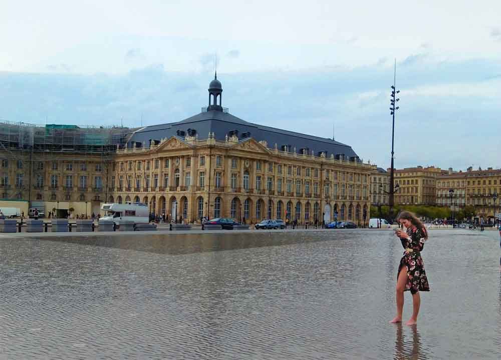 A woman standing in a pool of water while barefoot in front of the Palais de la Bourse in Bordeaux, France. The building is reflected in the water, and the woman is looking at the camera.
