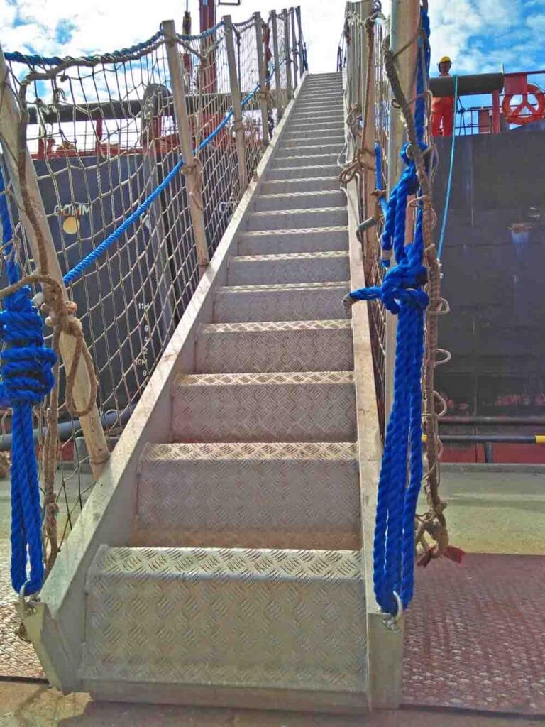 Ship's gangway with ropes and safety net rigged to the shore of a tanker terminal while the watchman stands near it on deck.