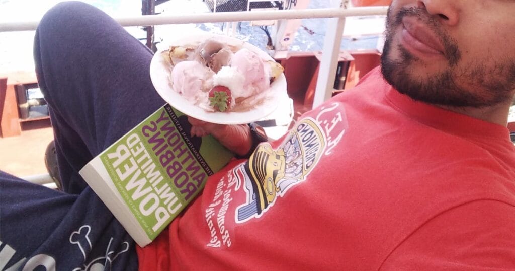 A seafarer holding an ice cream and a book that he's reading outside the ship.
