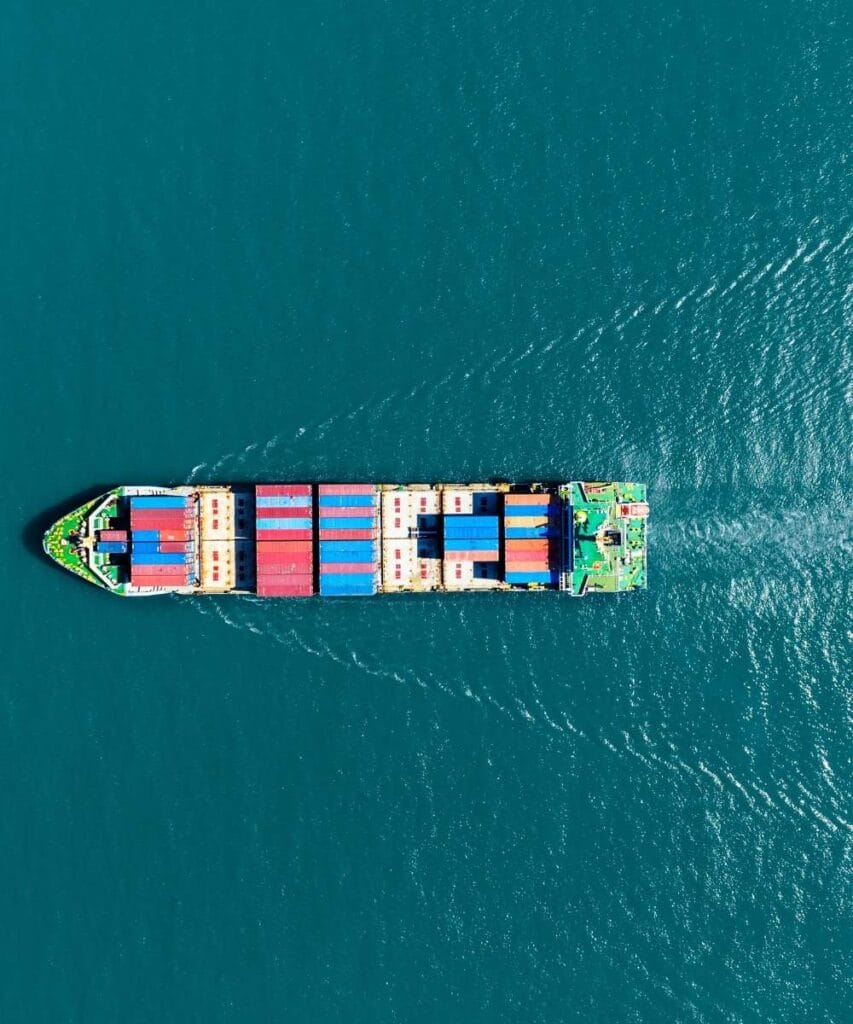A container vessel navigating in the ocean showing its wake and colorful cargoes on deck.